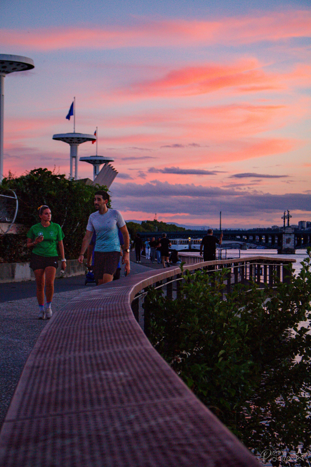 Deux joggers courent sur une promenade pavée en bord de fleuve au coucher de soleil, avec des structures futuristes et un pont en arrière-plan