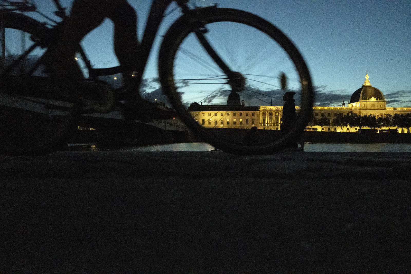 Vue nocturne à travers un cadre de roue de vélo, montrant un édifice illuminé avec dôme doré au bord d'une rivière au crépuscule.