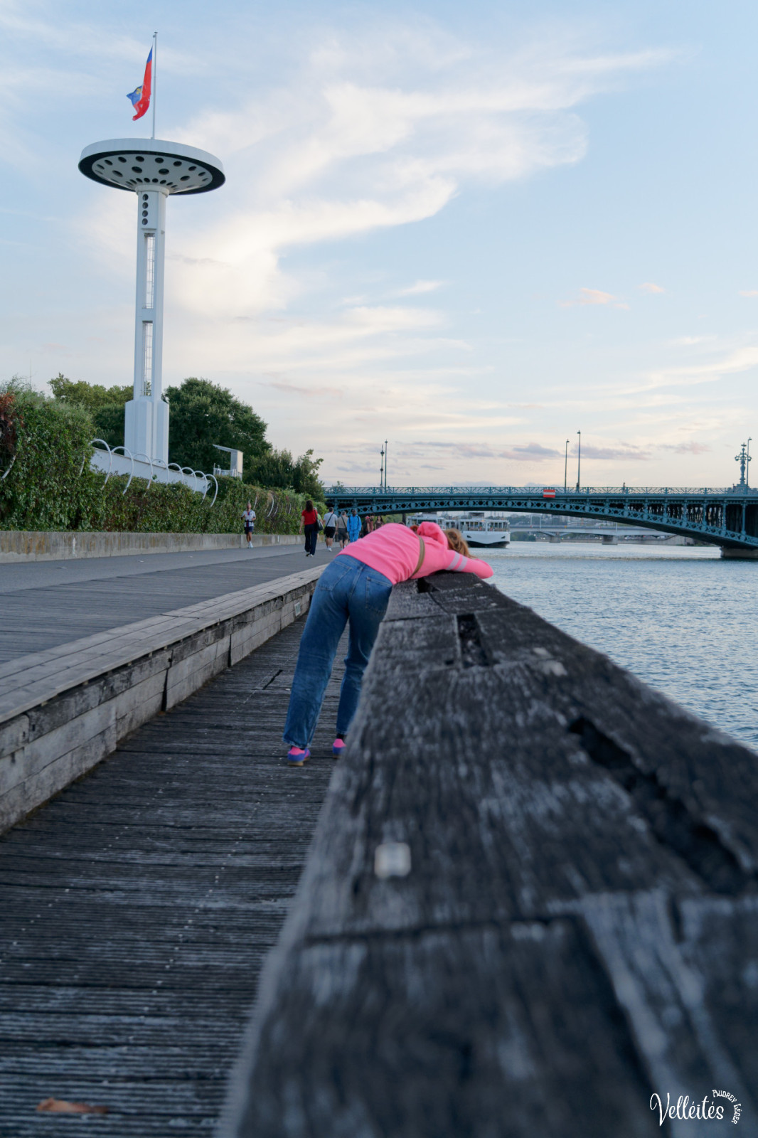 Une personne en vêtements roses et bleus fait de l'exercice sur une promenade côtière en bois, avec une tour-phare blanche, un pont et un fleuve en arrière-plan.