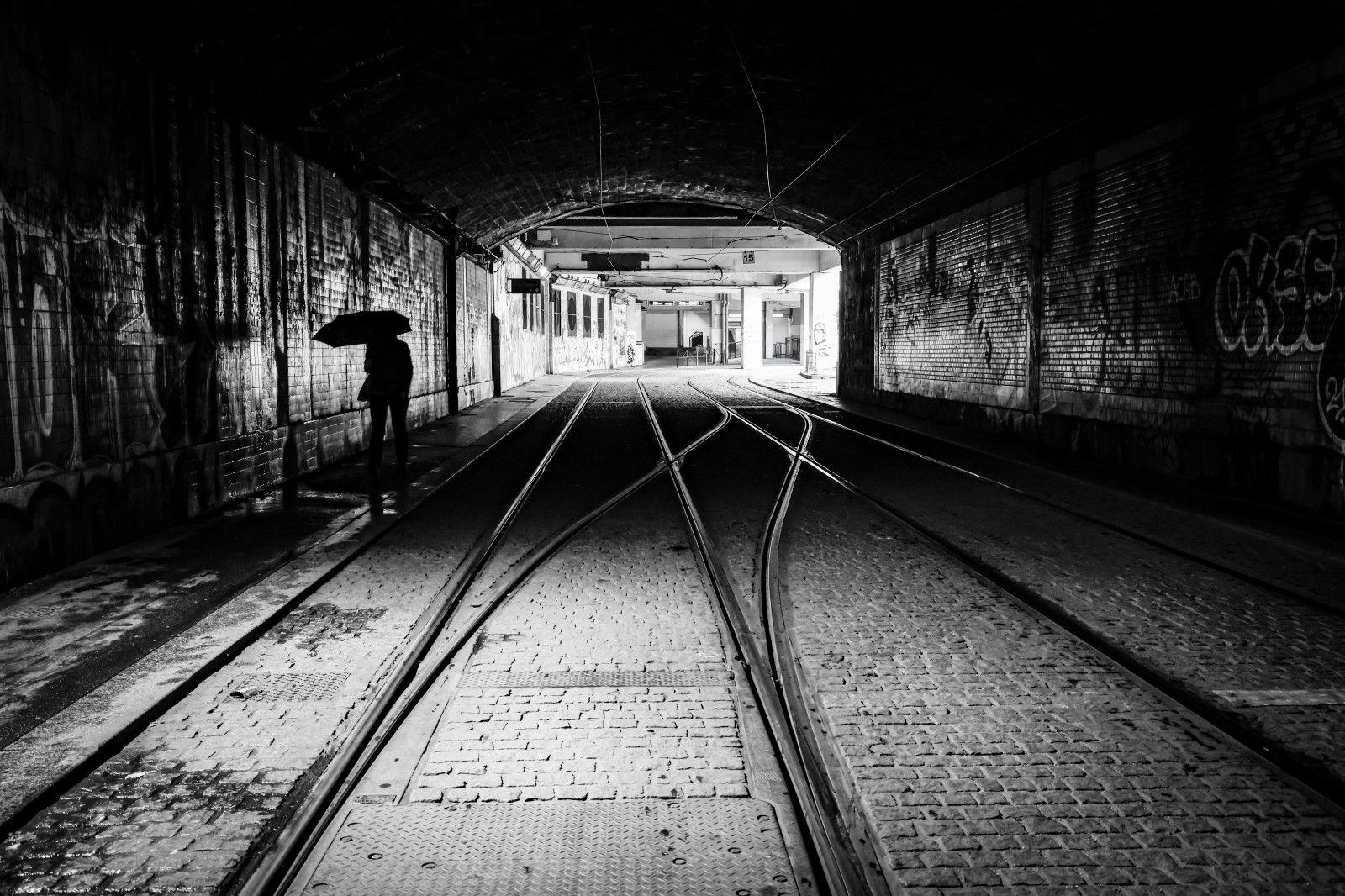 Silhouette solitaire sous son parapluie et les rails qui se croisent entre l’obscurité du tunnel et la lumière du dehors …